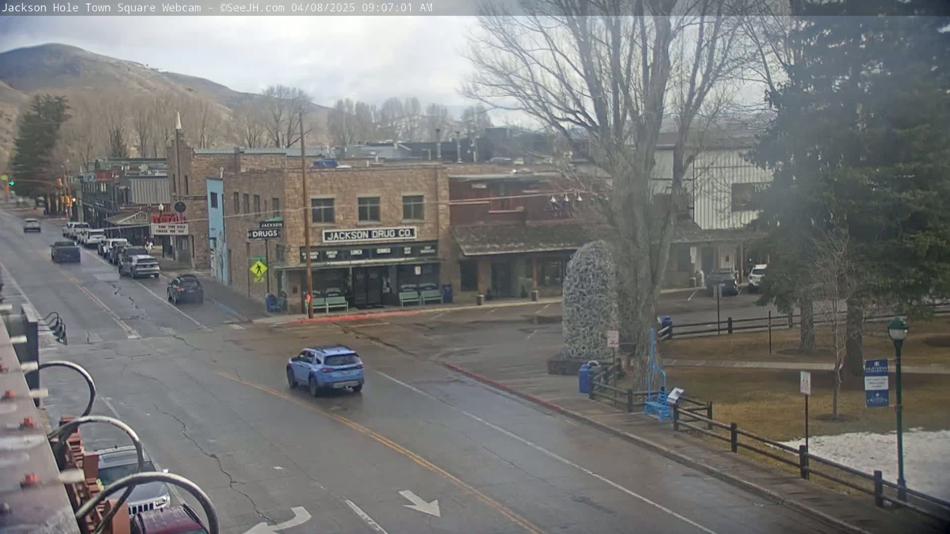 A mostly cloudy day reveals a town square with several cars on a street in front of buildings, a park with a few trees and some snow on the ground, and a mountain in the background.