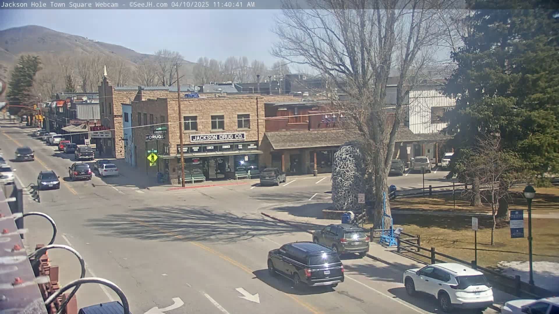 A sunny day in a town square, showing several cars driving on roads alongside buildings and a small park.