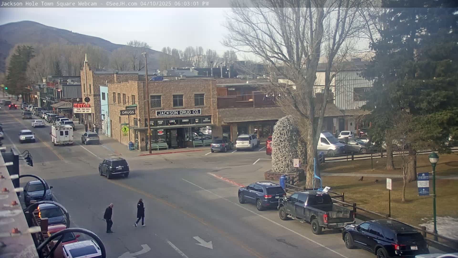 A partly sunny day reveals a town square with parked and moving vehicles, pedestrians, and buildings, including a drug store, set against a backdrop of mountains.