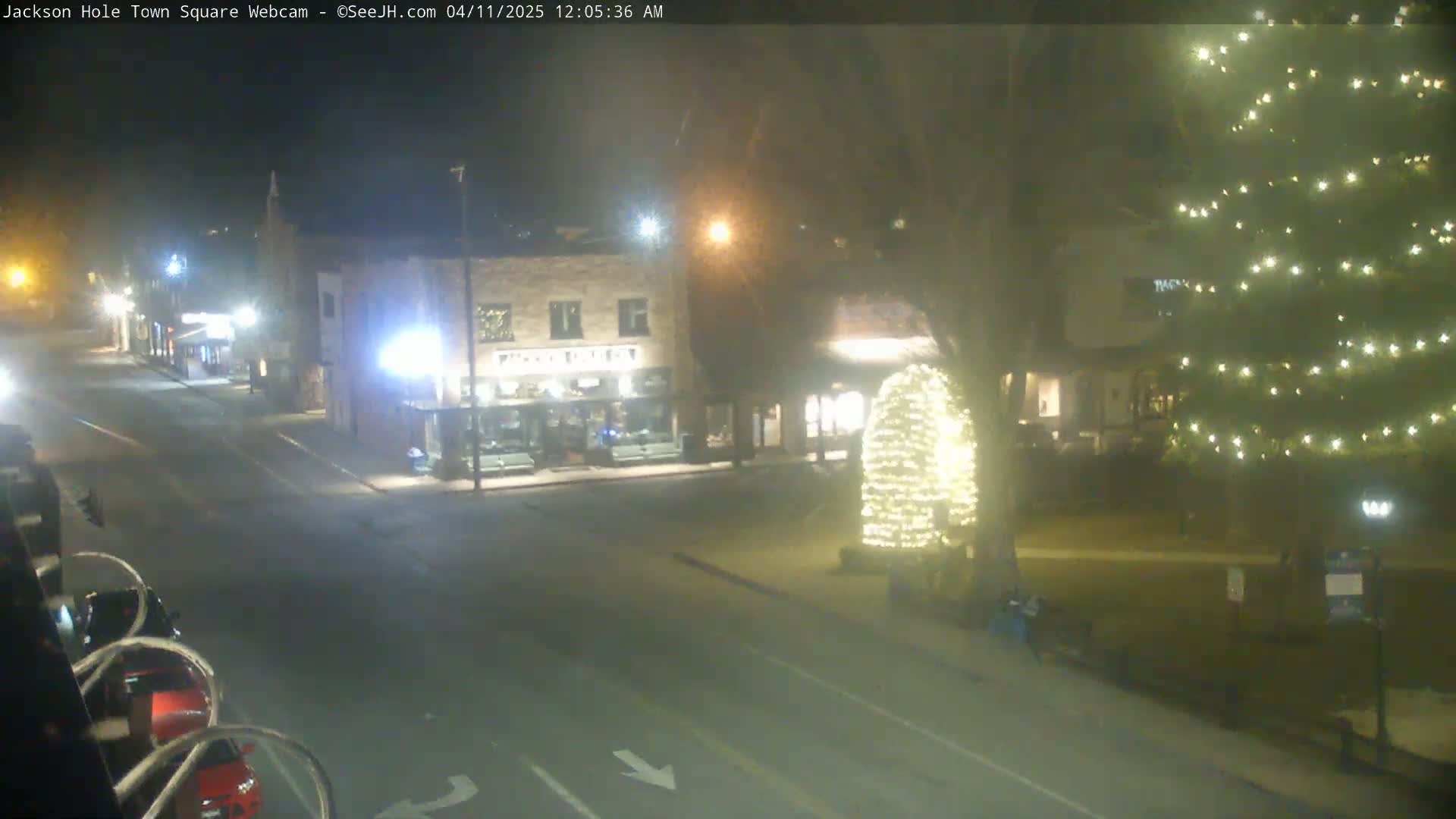 A nighttime view of a town square with a street, buildings, and a Christmas tree illuminated with lights.