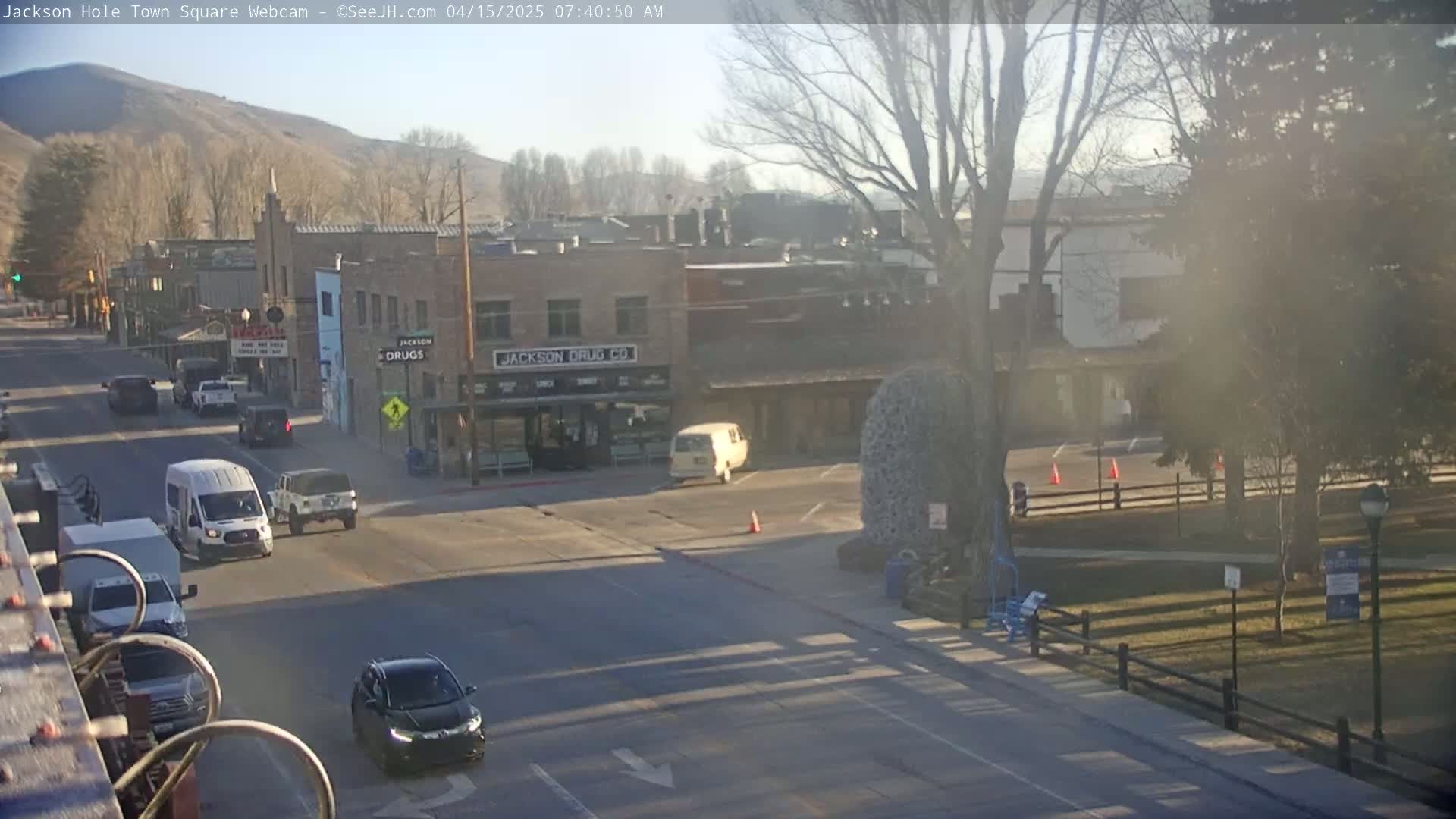 A mostly sunny morning view of a town square with several vehicles at an intersection, buildings, and a park in the background.