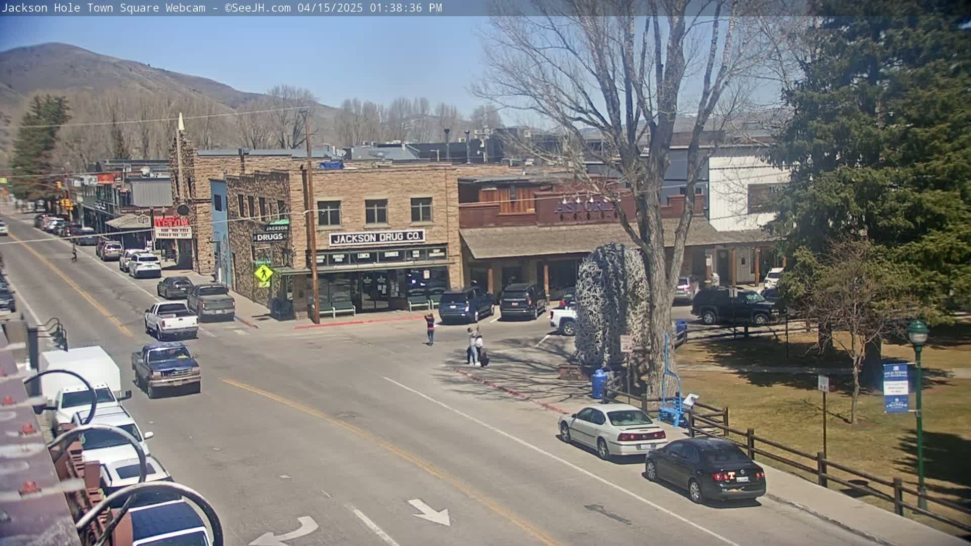 A sunny day in a town square shows several cars parked and driving along streets, with buildings and a park visible.