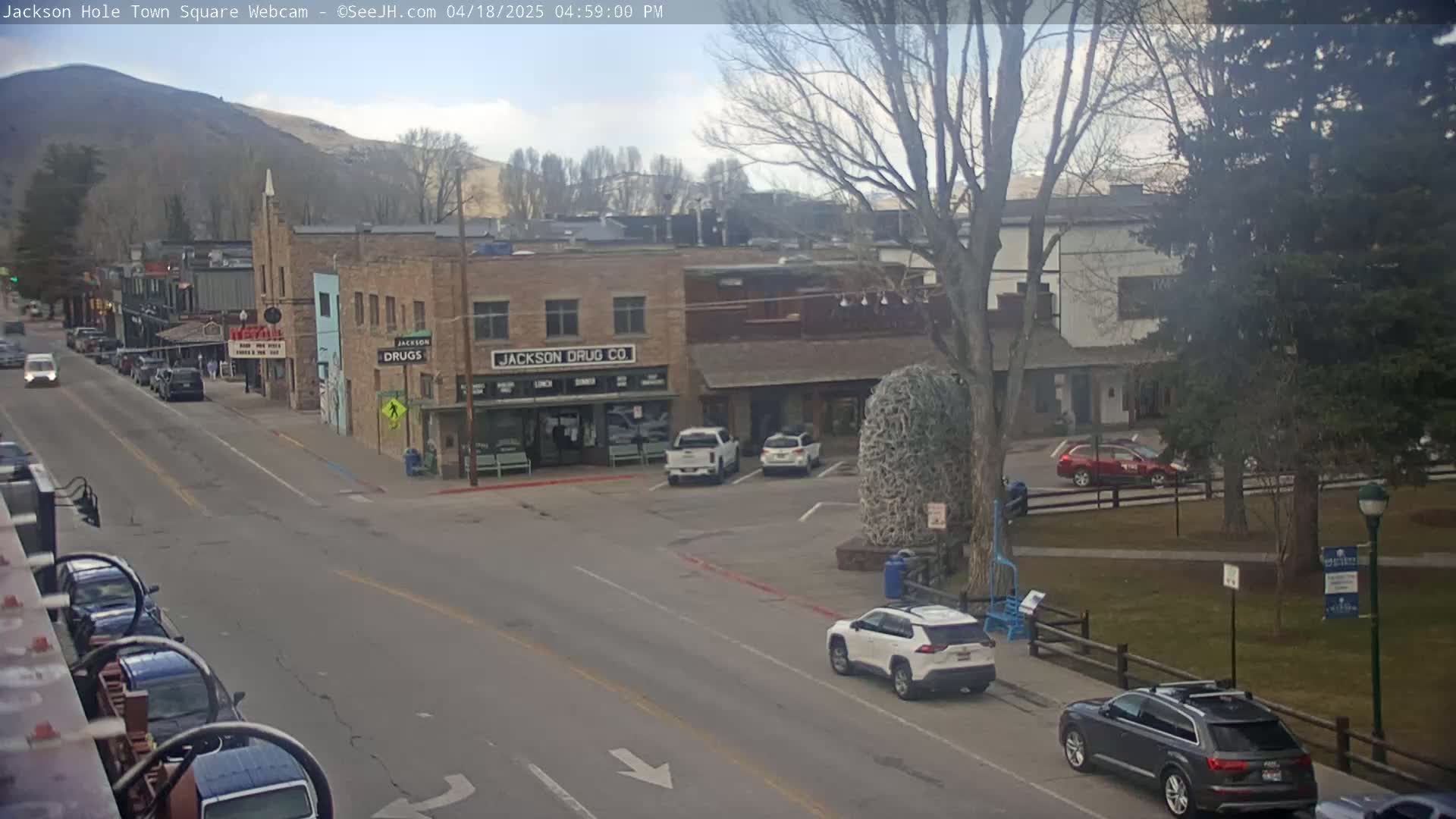 A partly cloudy day in a town square shows several cars parked and driving, brick buildings, and leafless trees.