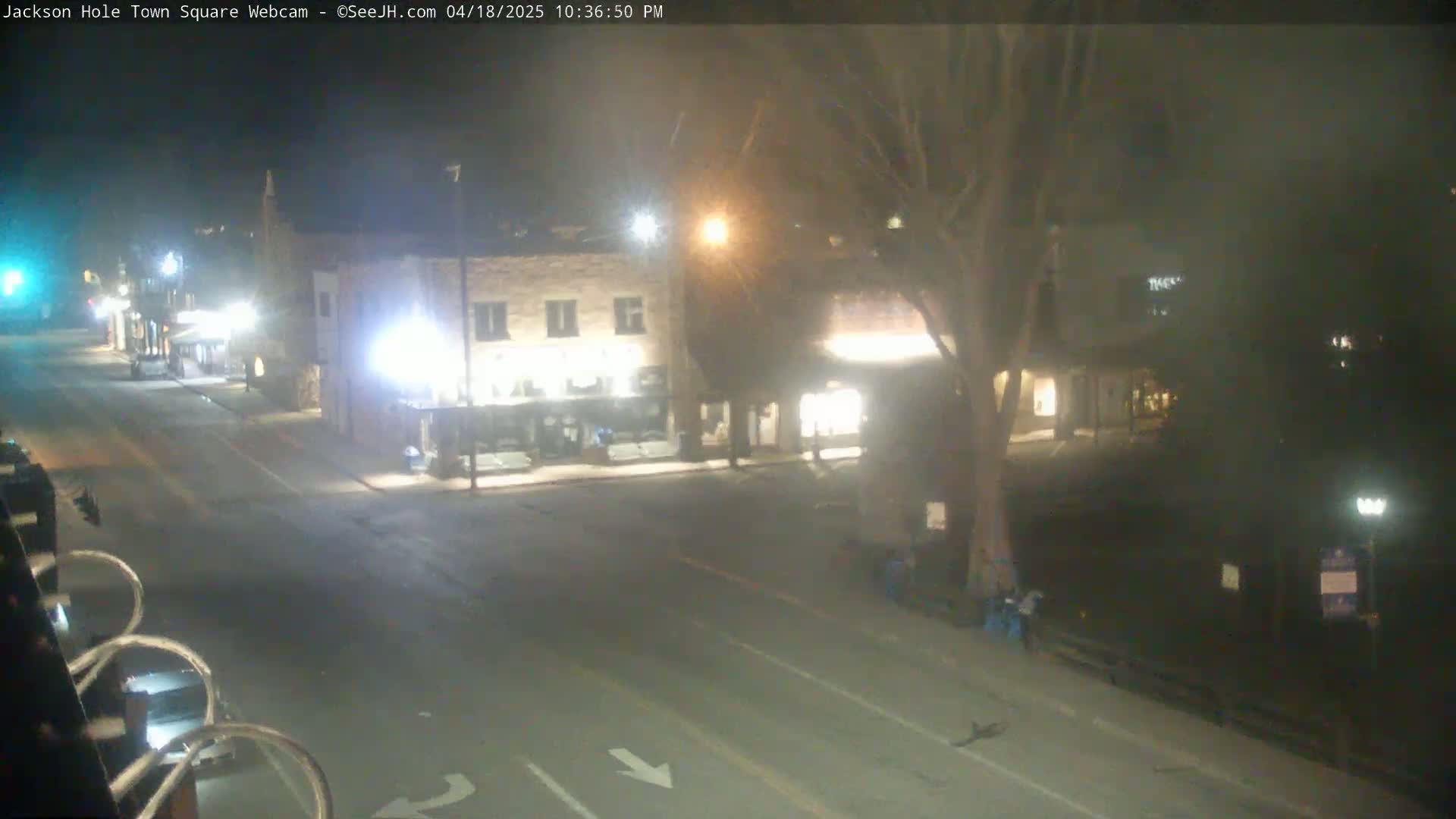 A nighttime view of a mostly empty town square with a few buildings and a large tree, under hazy conditions.