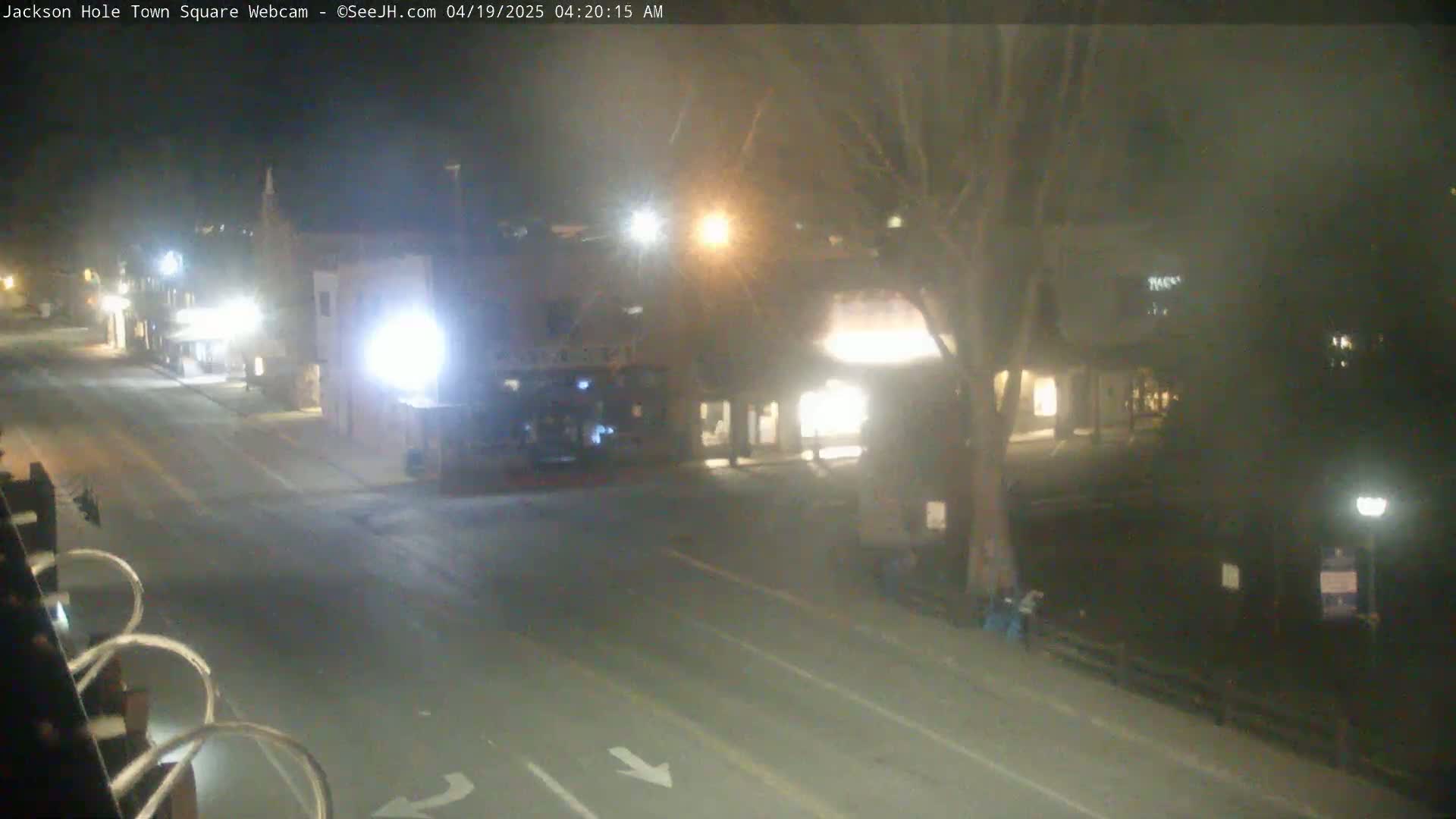A nighttime view of a mostly empty town square, with several buildings lit and a street with lane markings.