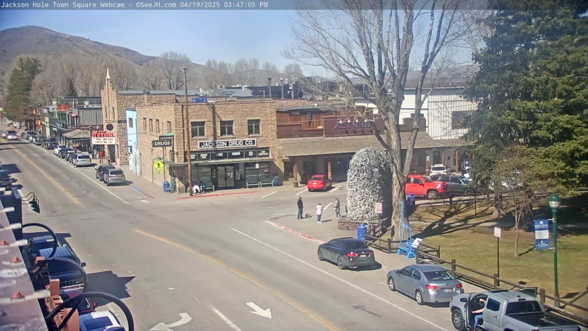 A sunny day in a town square shows several cars parked and driving, with buildings and trees surrounding the area.