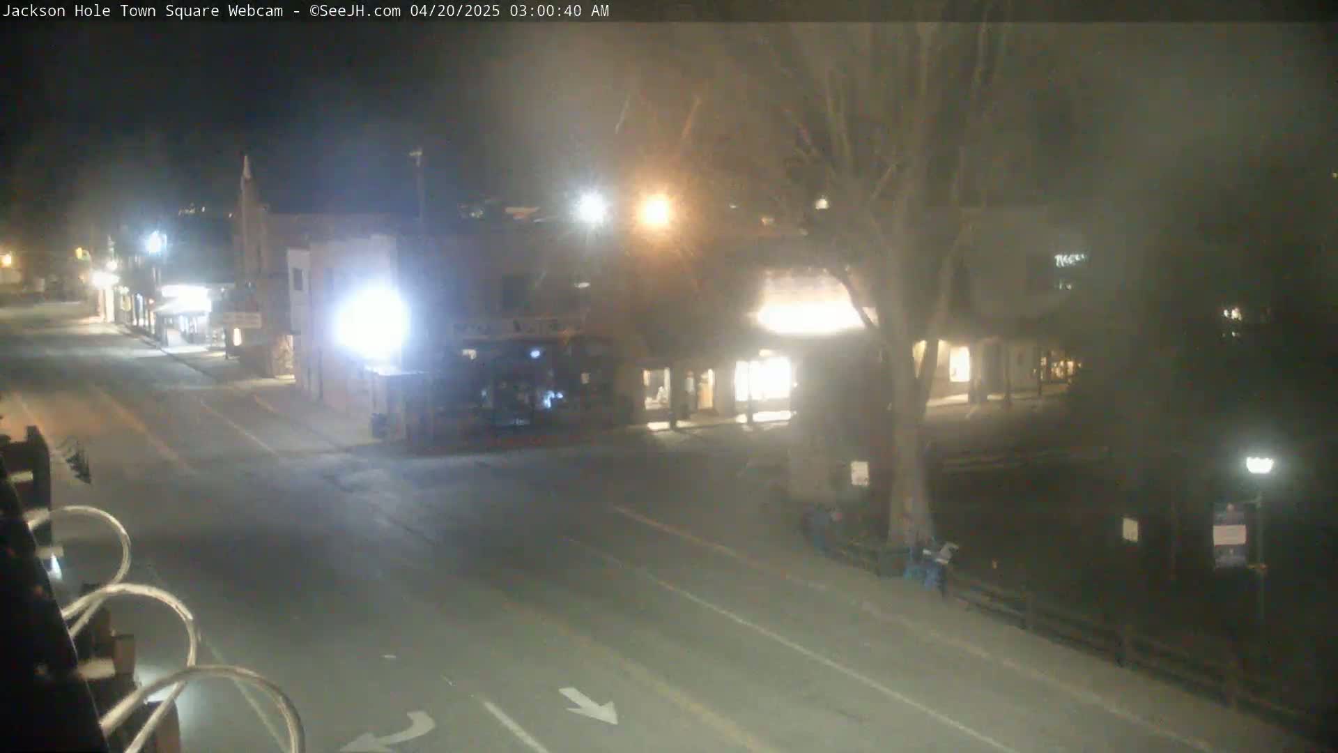 A nighttime view of a mostly empty town square with streetlights illuminating a quiet road and nearby buildings.