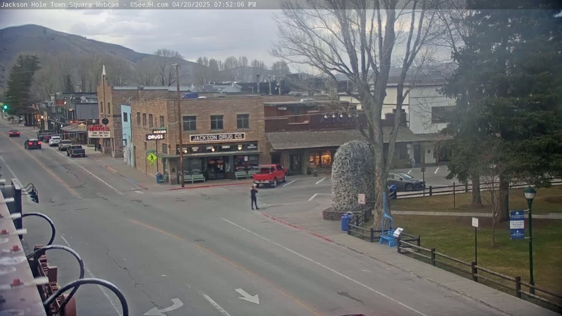 A mostly empty town square with a few cars and a person walking, under an overcast sky.