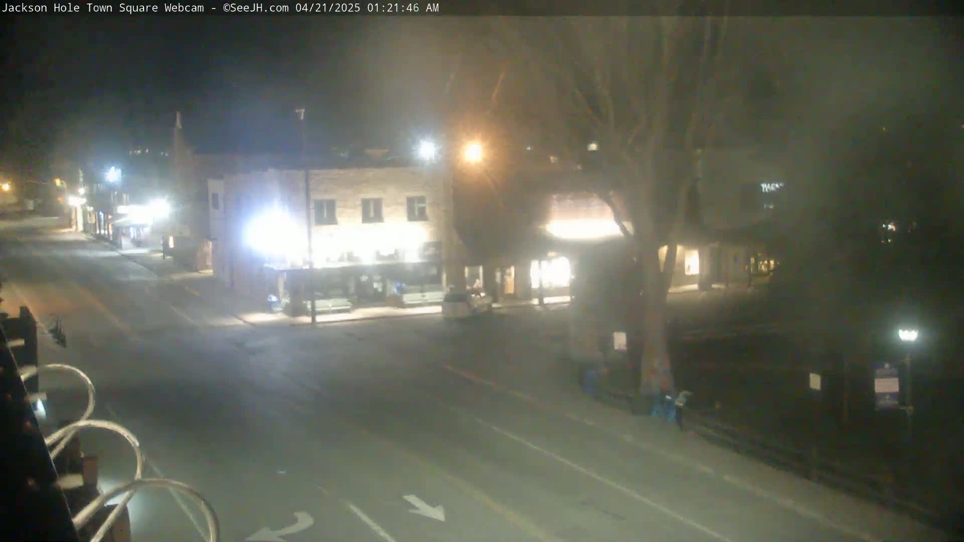 A nighttime, mostly empty street scene shows a few buildings with illuminated windows, a lone vehicle, and a tree in the middle of a town square.