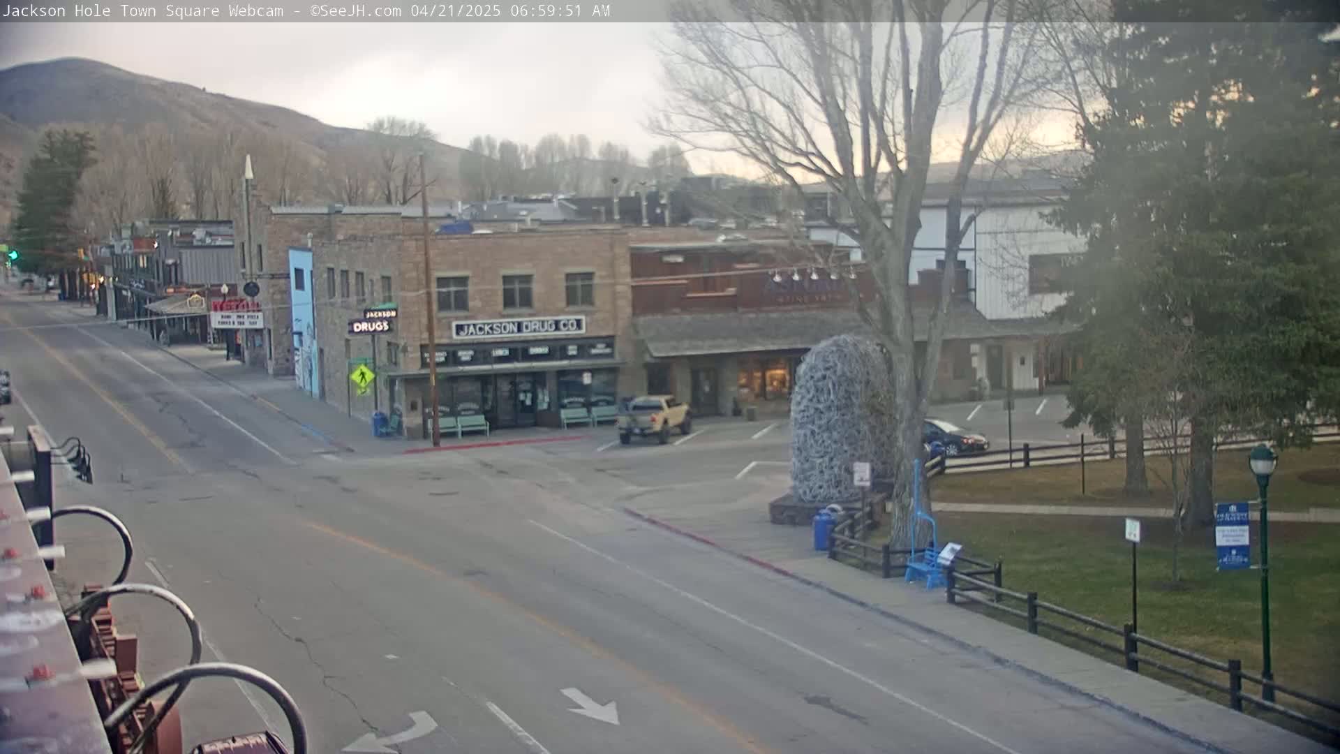 A mostly sunny town square with a few cars parked and driving, trees, and buildings, one of which is a drugstore.
