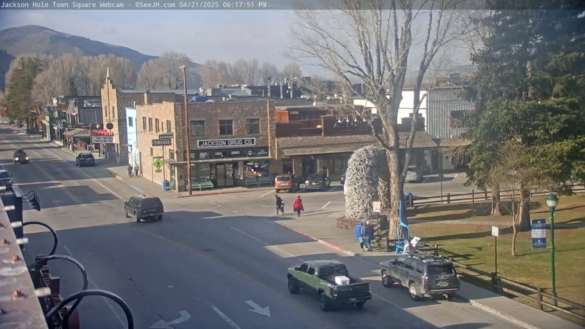 Under a partly sunny sky, vehicles are parked and driving through a town square, past buildings and a grassy area with trees.