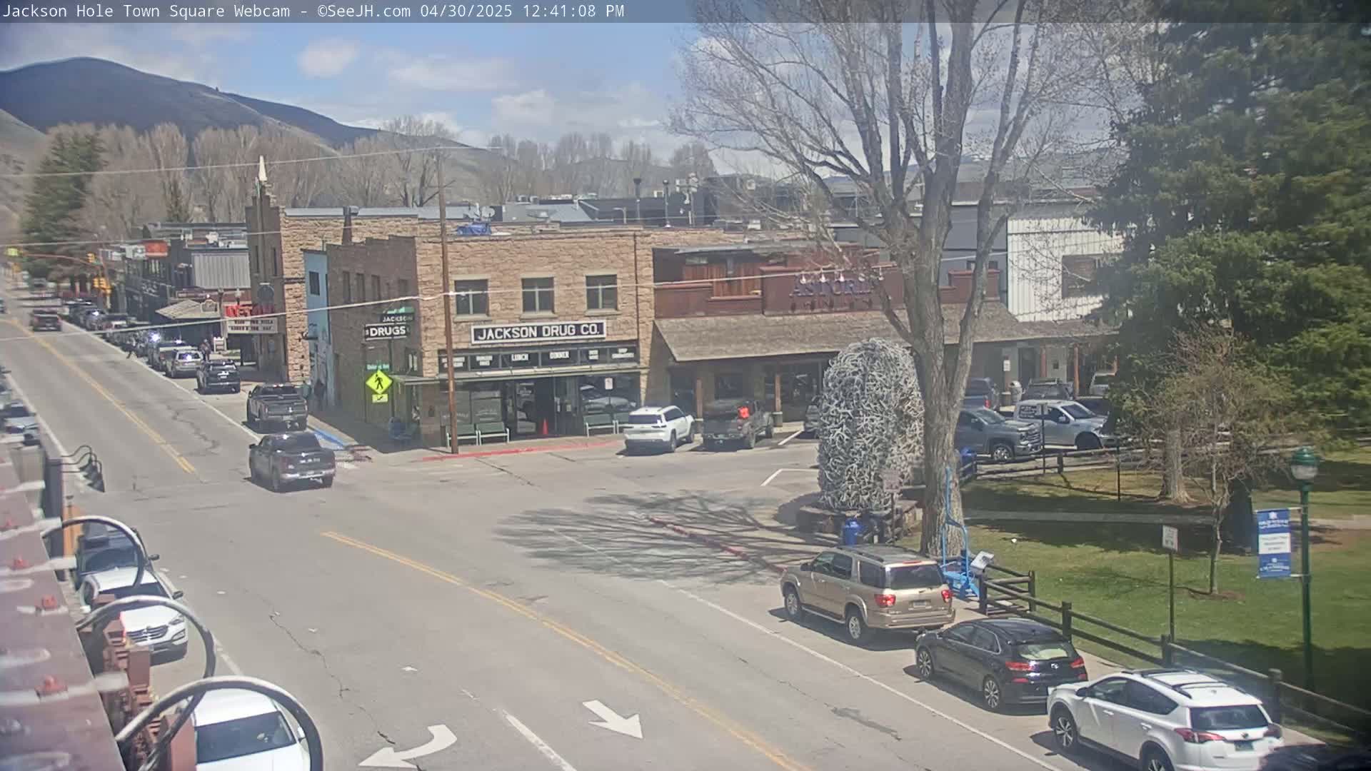 A sunny day in a town square, showing several cars parked and driving around buildings, with a tree in the center of the square and mountains in the background.