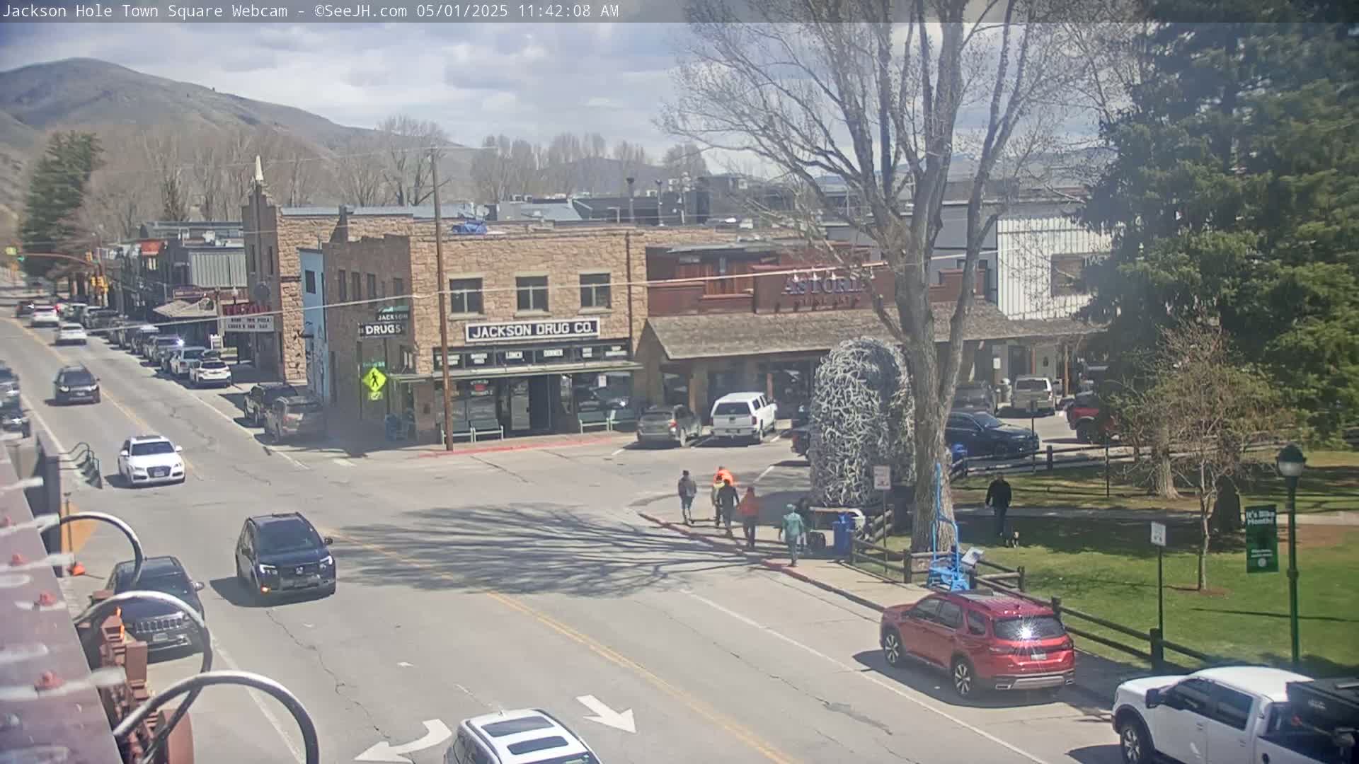 A partly sunny day reveals a town square with cars driving on streets, pedestrians walking, and buildings lining the roads; trees and a grassy park are visible.