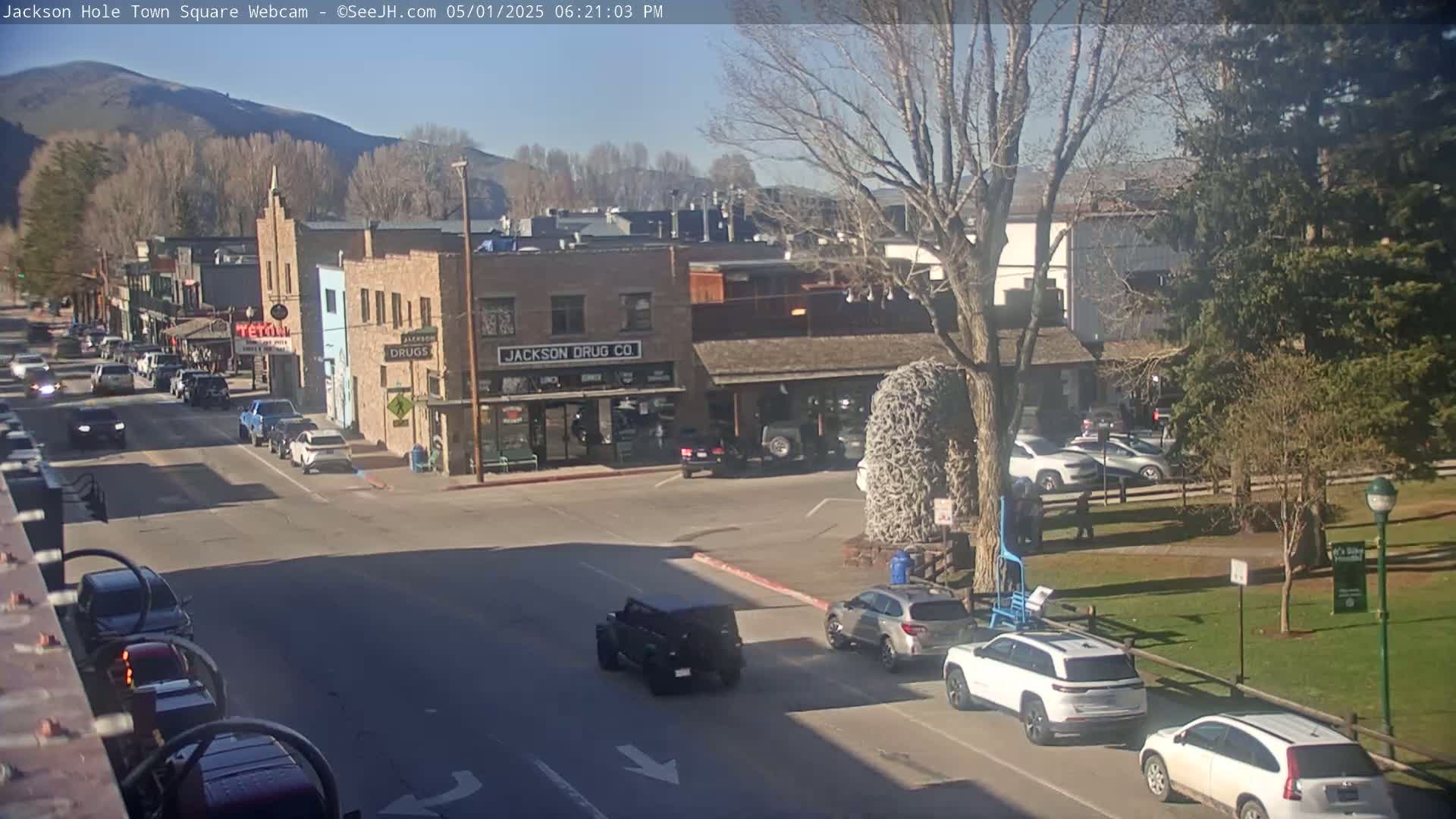 A sunny town square, with cars parked along the street and several vehicles driving, sits under a mostly clear sky with bare trees and distant mountains.
