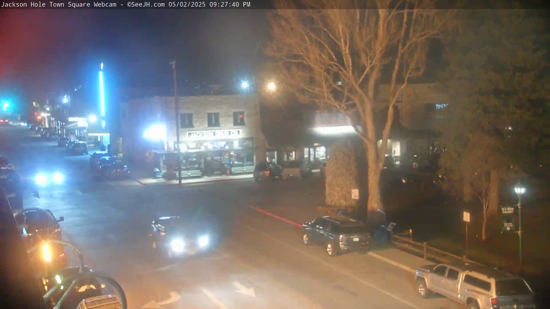 A nighttime view of a town square with several cars on the road and parked, under a clear sky.