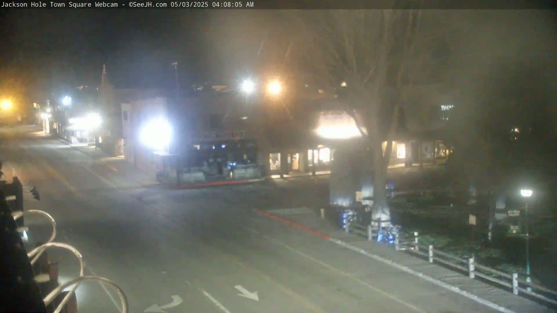 A nighttime view of a mostly empty town square with several buildings and a vehicle, under hazy conditions.