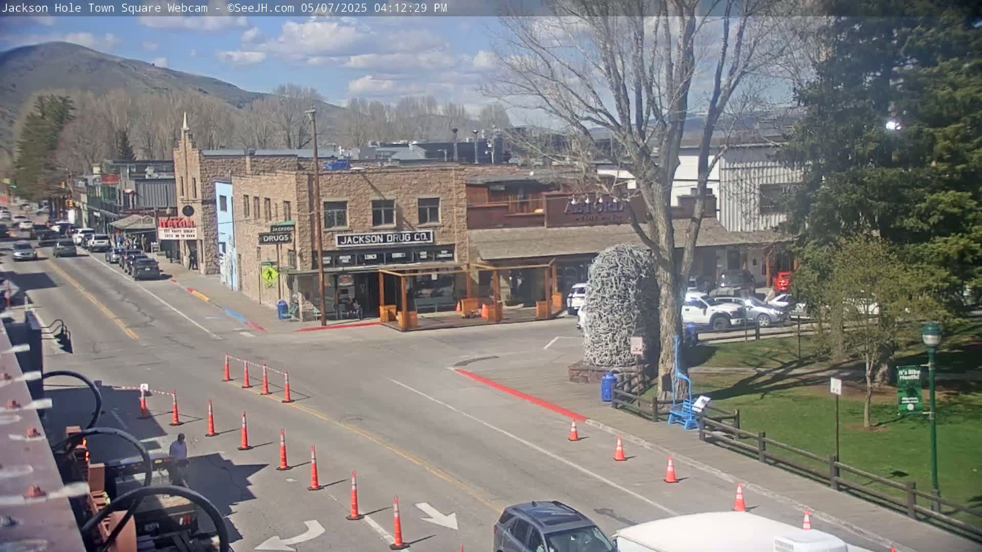 A sunny day reveals a town square with stone buildings, parked cars, and a street with orange traffic cones, bordered by grass and trees under a mostly clear sky.