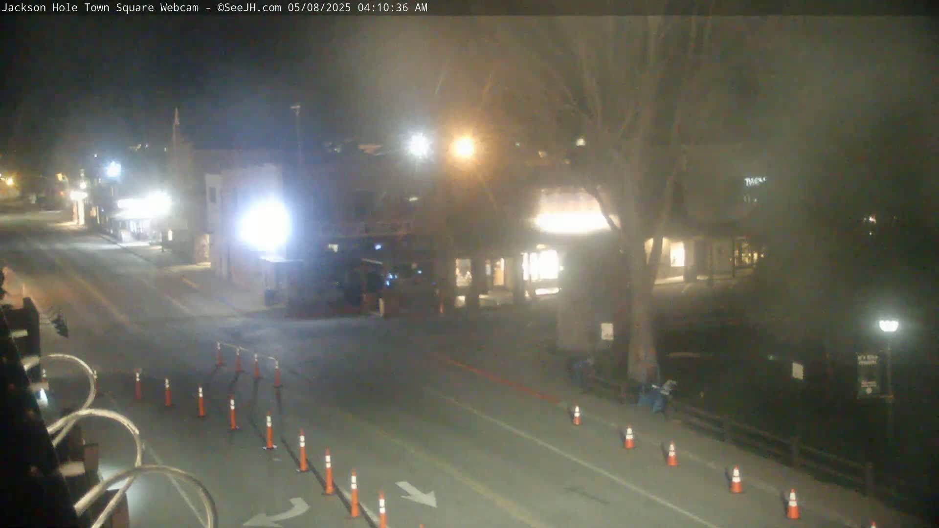 A nighttime, slightly hazy view of a town square with a street, buildings with lights on, and orange traffic cones lining a roadway.