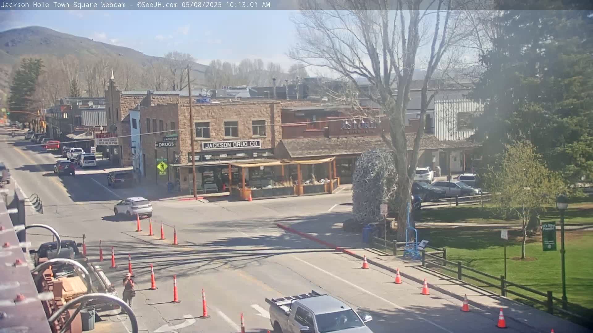 A sunny town square scene shows a street intersection with cars, construction cones, and buildings, including one labeled "Jackson Drug Co.", with a park and trees in the background.