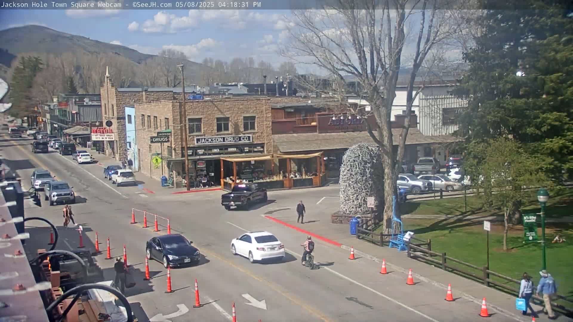 A sunny day in a town square shows several cars driving on the street, pedestrians walking on the sidewalk, and a person riding a bicycle, all in front of buildings and a grassy park.