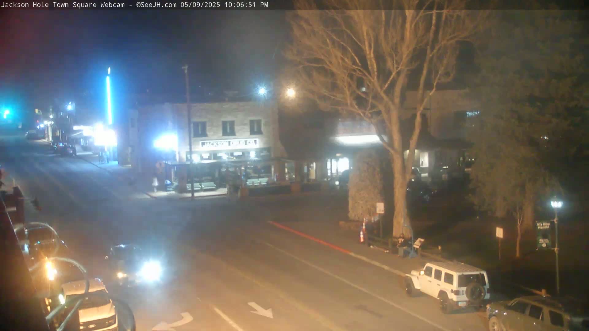 A nighttime, dimly lit town square shows several vehicles parked and driving along a road, with some people standing near buildings under a mostly bare tree.