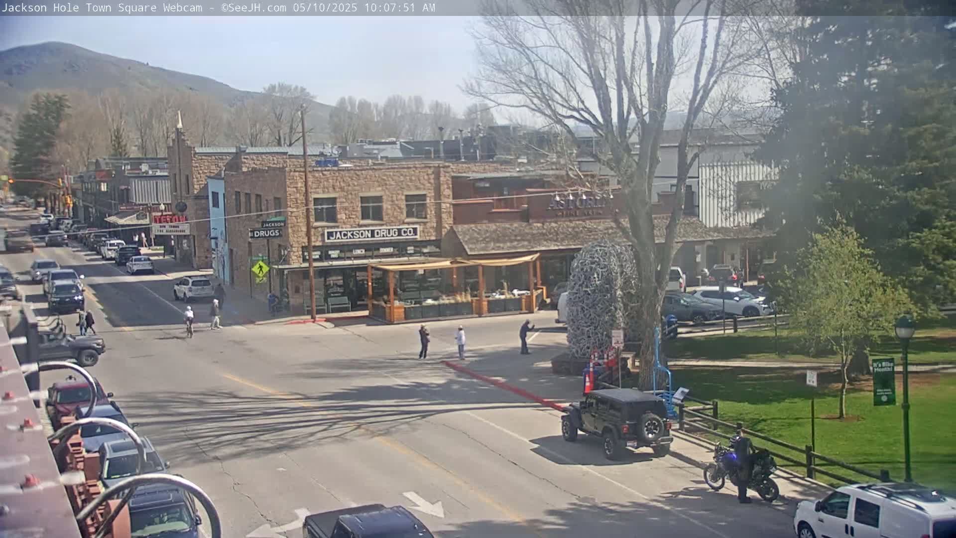 A sunny day in a town square shows several parked cars, pedestrians, and bicyclists, with buildings lining the street and a grassy park area nearby.