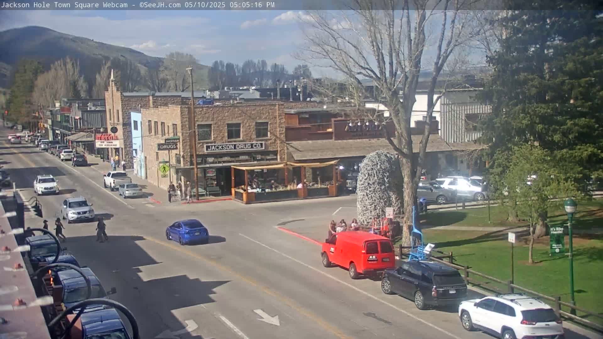 A sunny town square scene shows cars driving on a street lined with buildings, including one with a visible sign that says "Jackson Drug Co.", under a mostly clear sky.