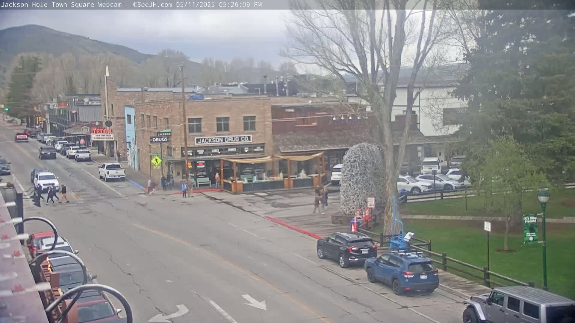 A partly cloudy day shows a town square with several cars parked and driving, pedestrians walking, and buildings lining the street, one of which is a drug store.
