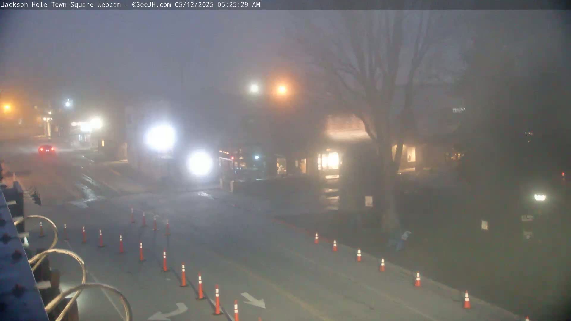 A foggy street scene at night shows a road with orange traffic cones, a few vehicles, and buildings barely visible in the distance.