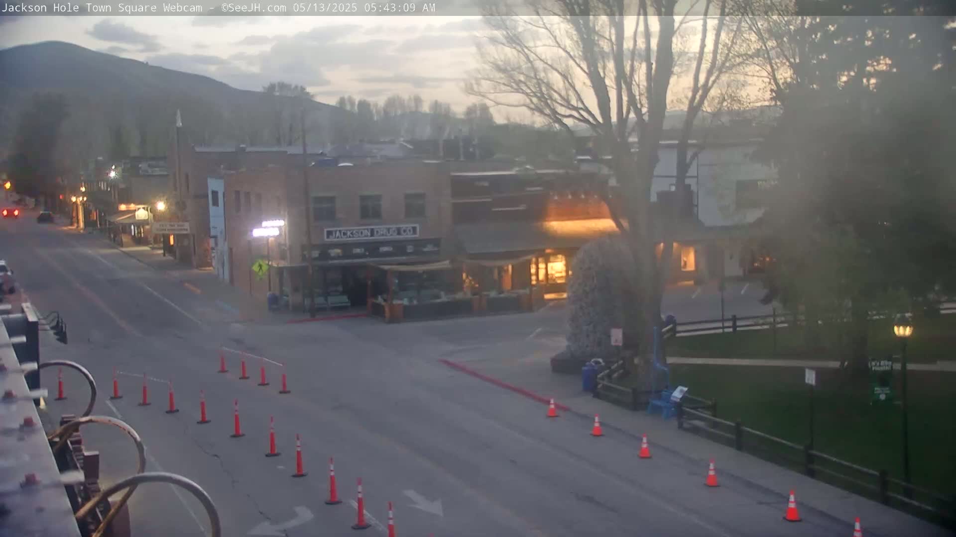 A hazy dawn reveals a mostly empty town square with several buildings, some trees, and orange traffic cones lining a street.