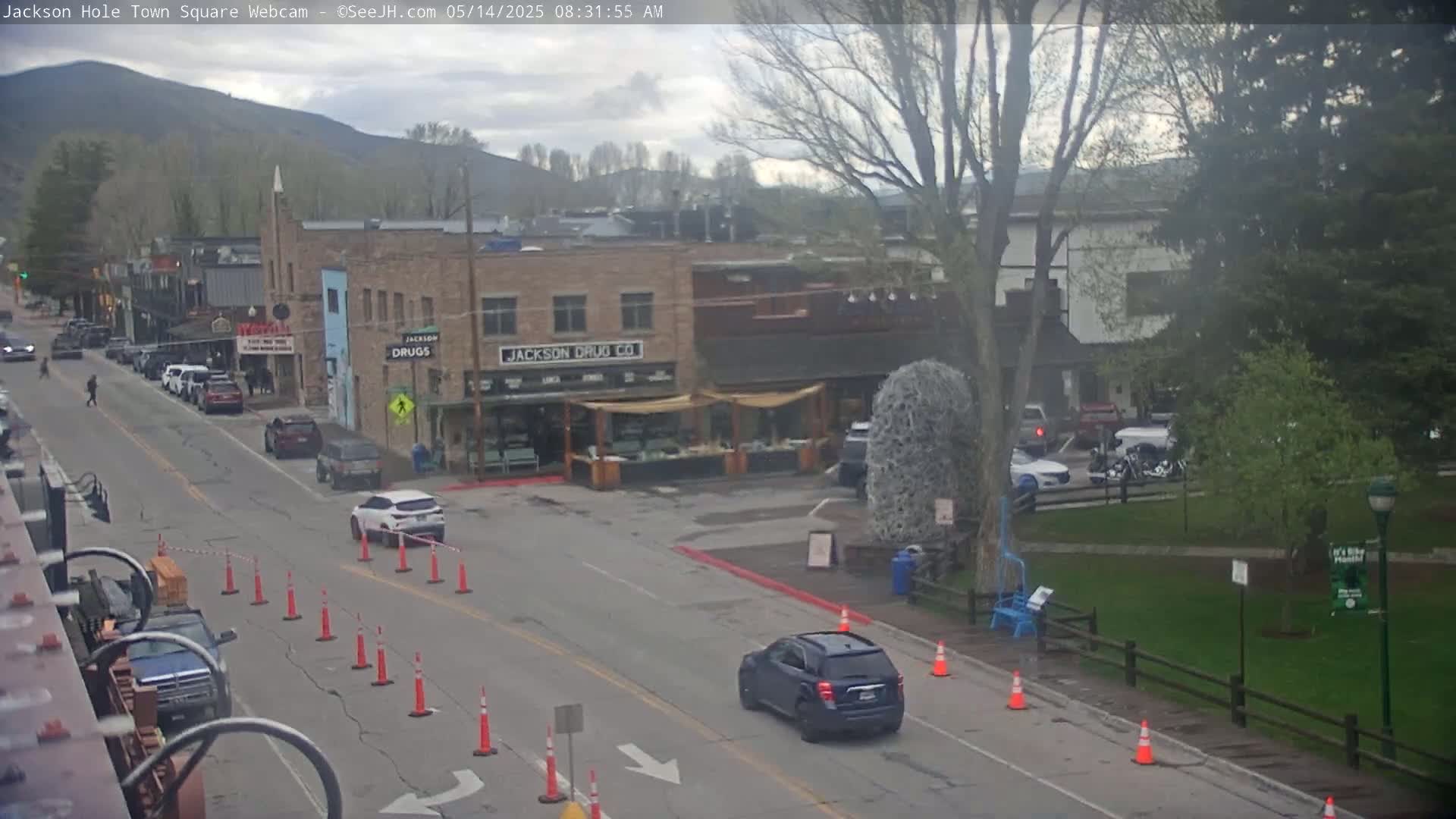 A partly cloudy day reveals a town square with several cars parked along the street, in front of shops and buildings, with orange traffic cones marking a section of the road.