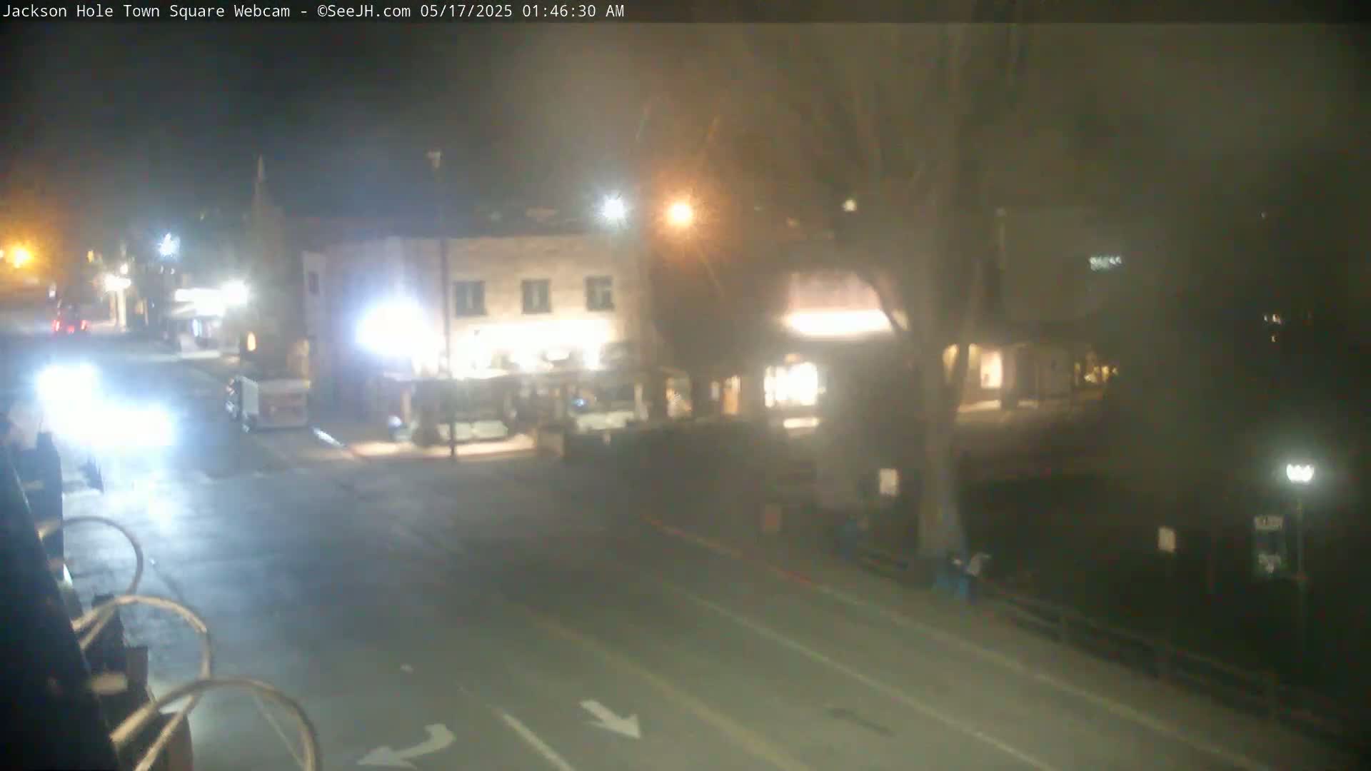 A nighttime view of a mostly empty town square with a few vehicles, some buildings with lights on, and a tree.