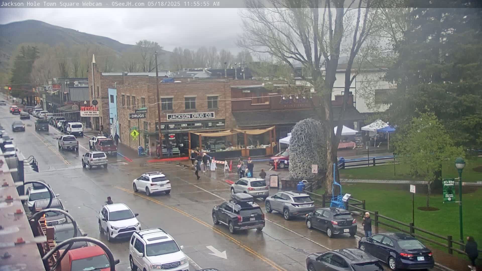A wet town square with cars parked along the street and people walking amidst buildings under an overcast sky.