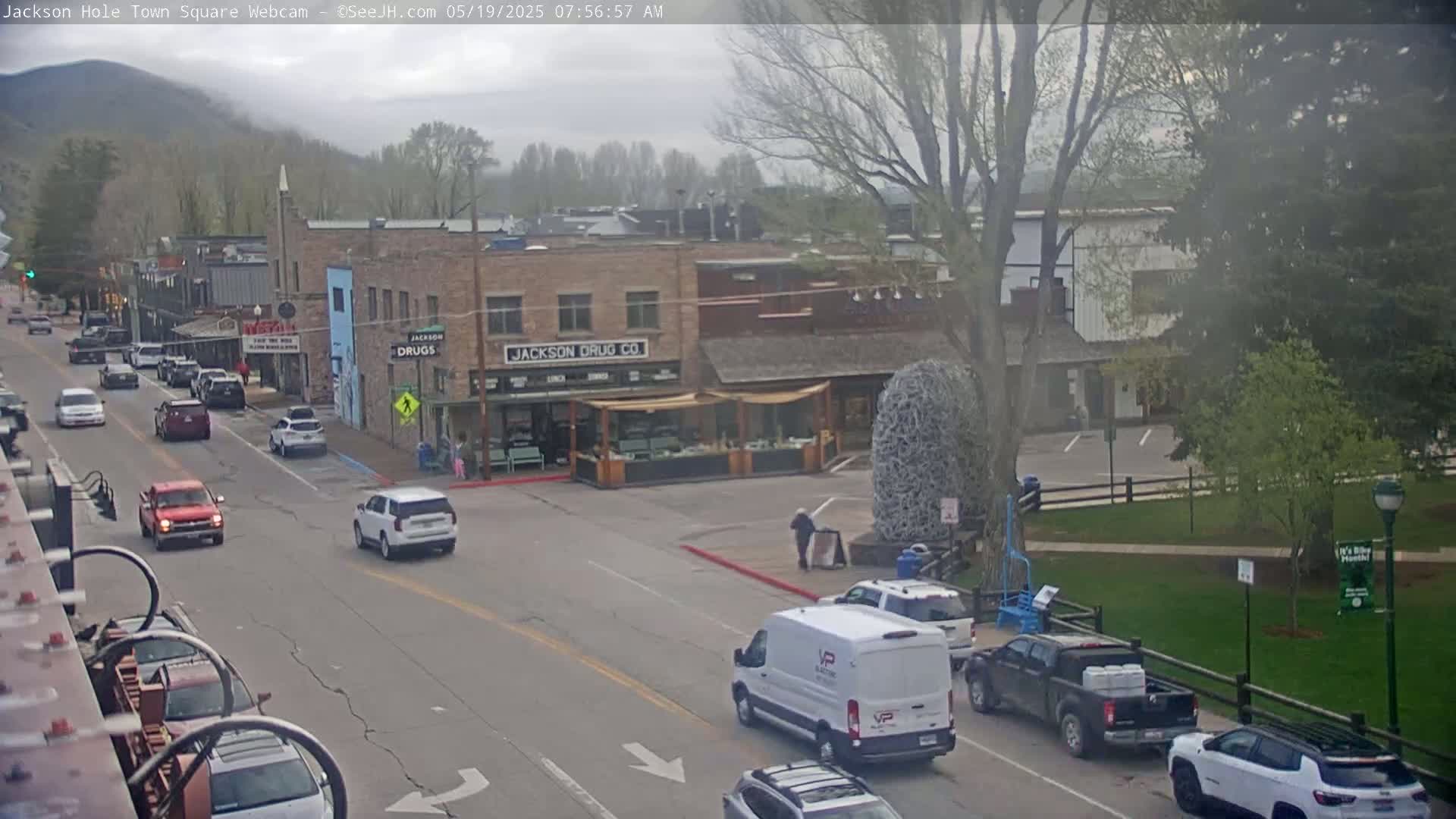 A mostly cloudy day in a town square with several vehicles on the street and a few pedestrians near buildings, one of which is a drugstore.