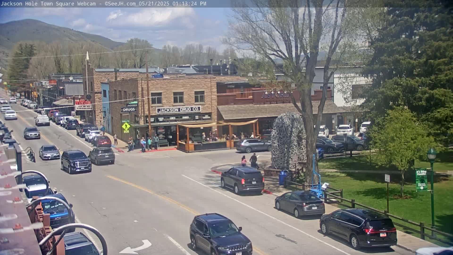 A sunny day in a town square, with cars parked and driving along a street lined with buildings, including one with a drugstore, and a grassy park area visible across the street.