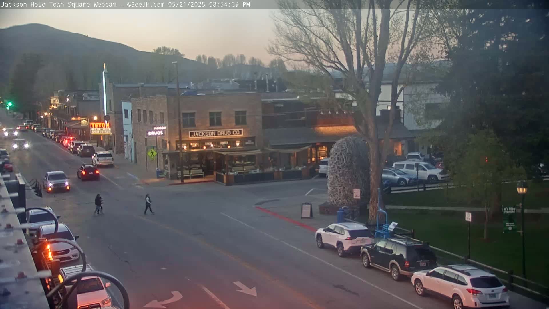 A street scene at dusk shows cars parked and driving along a road in front of buildings, with a few pedestrians crossing the street.