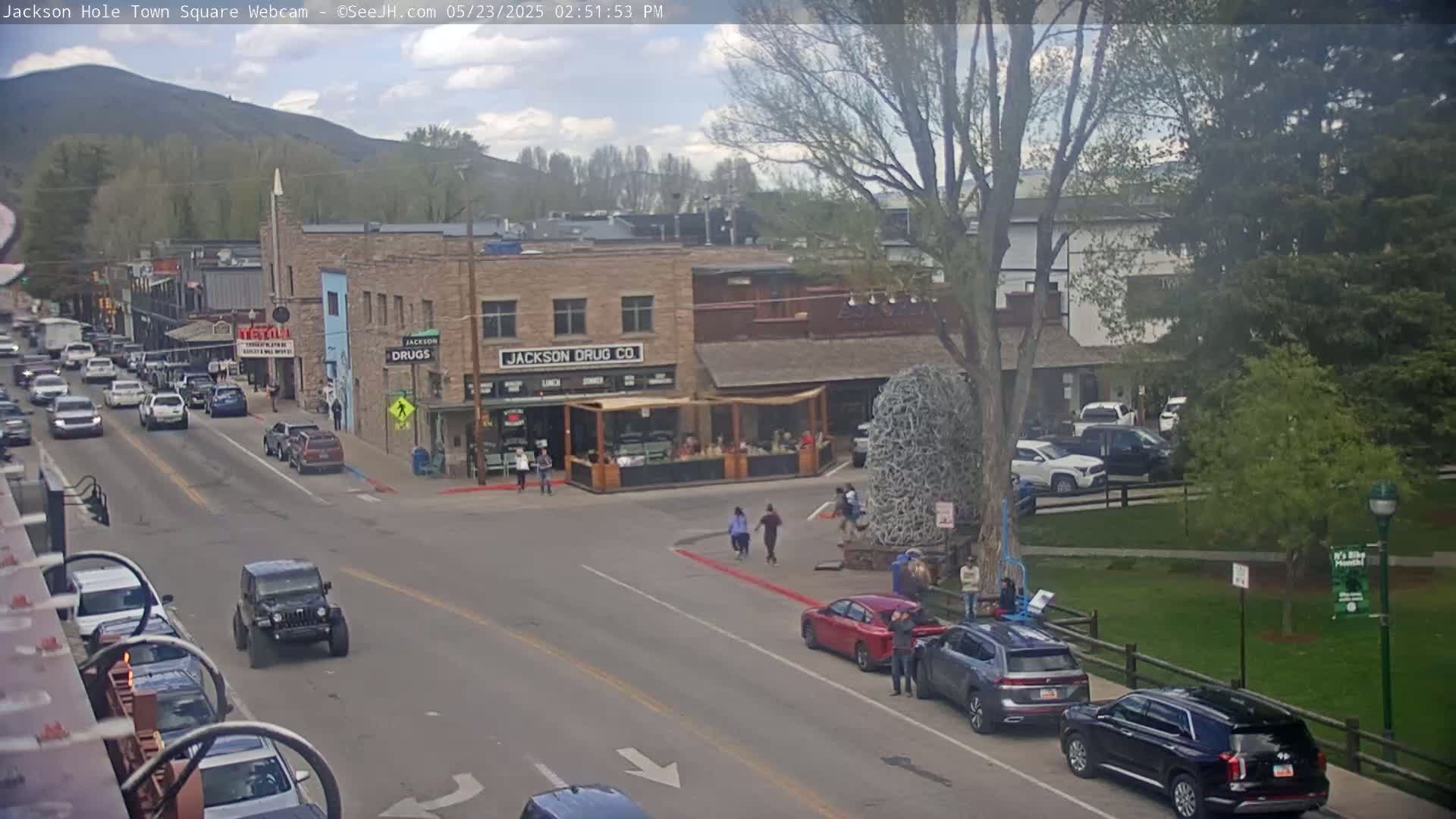 A partly sunny day in a town square shows several cars parked and driving along streets lined with buildings, with people walking in a nearby park.
