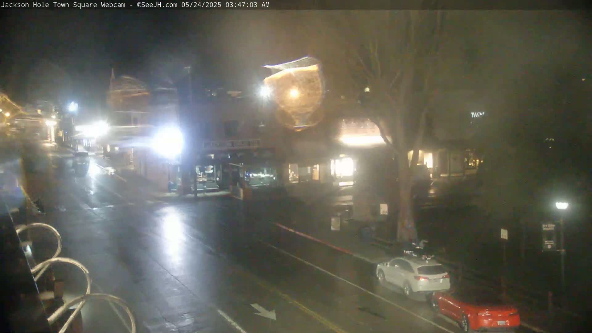 A wet, nighttime street scene shows a town square with several buildings, cars, and a tree, under dim light.