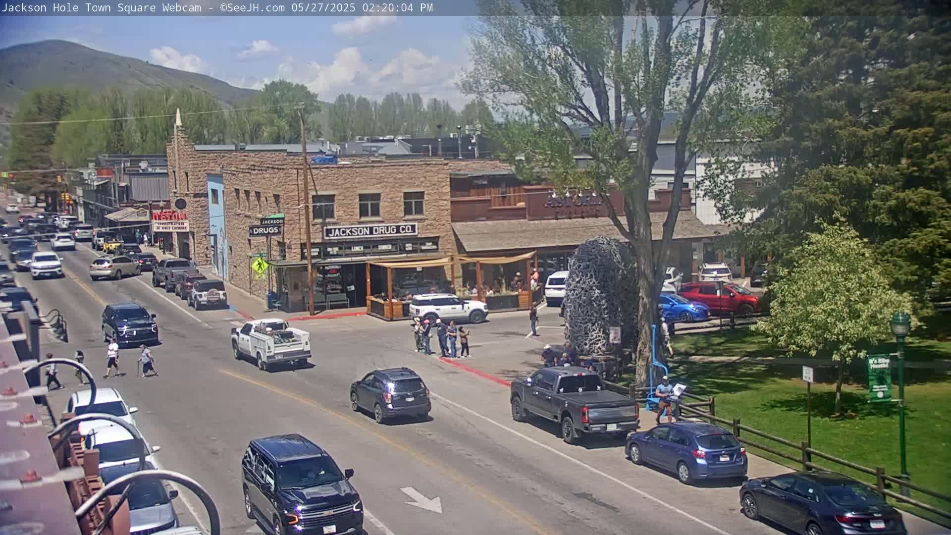A sunny day in a town square shows several cars driving on a street lined with buildings, some pedestrians crossing the street, and a park with lush green grass.