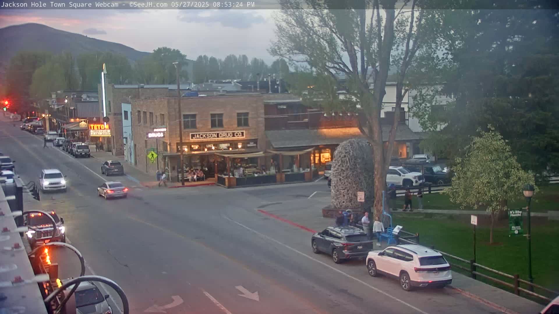 A town square at dusk, with cars parked along the street and several vehicles driving by, under a partly cloudy sky.