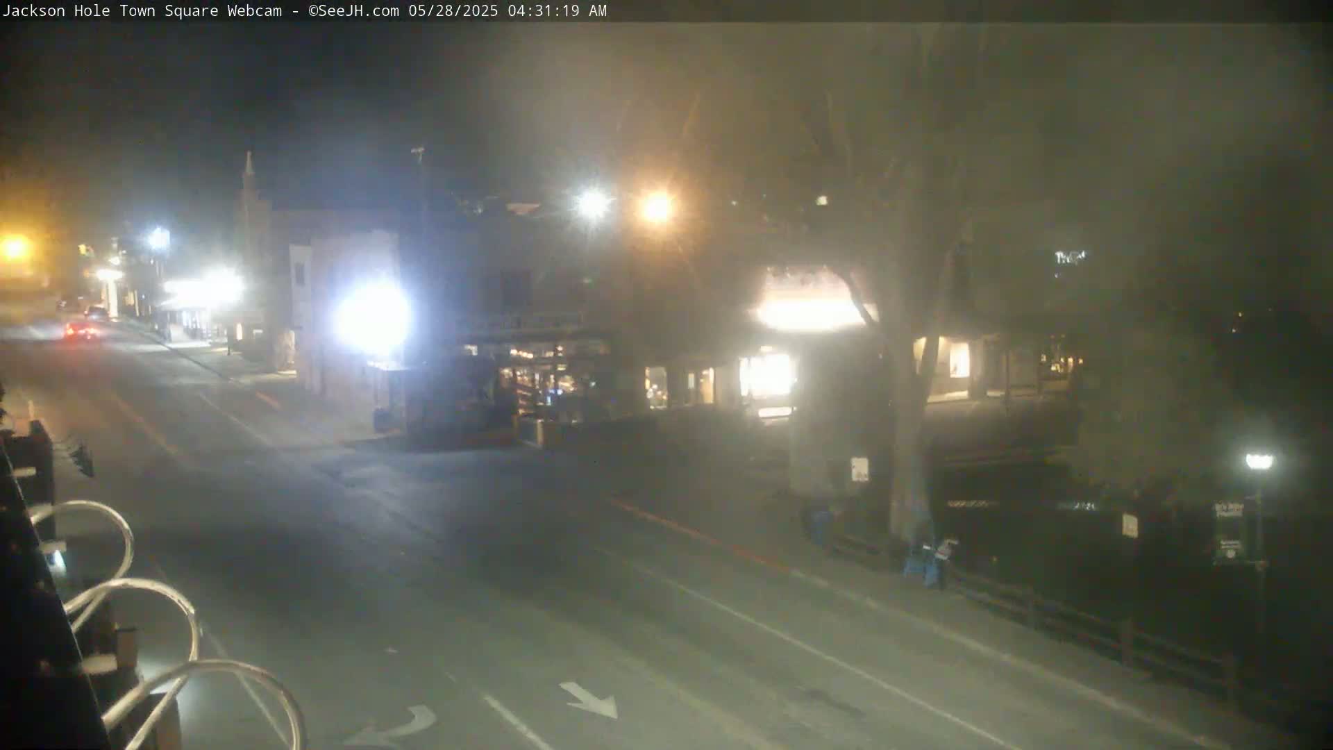 A mostly empty, fog-shrouded street at night shows a few cars and buildings with lights on.