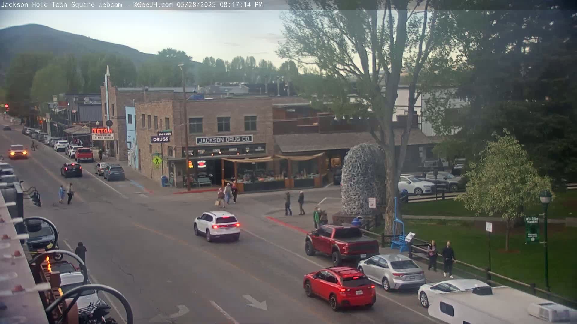 A street scene in a town square shows several cars on a street, pedestrians walking on sidewalks, and buildings, under partly cloudy, late afternoon light.