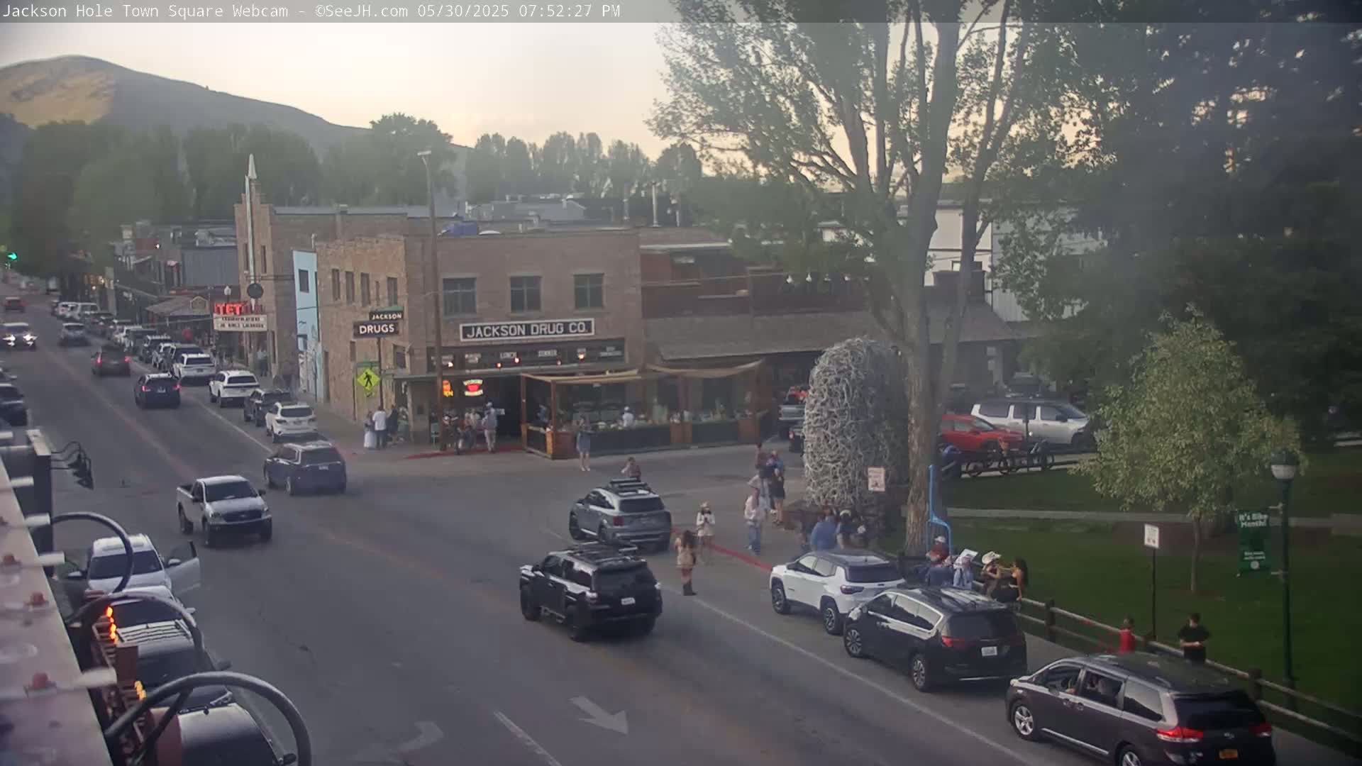 A street scene in a town square shows several cars parked and driving on a road lined with buildings, one of which is a drugstore, with people walking on the sidewalk under a hazy, late afternoon sky.