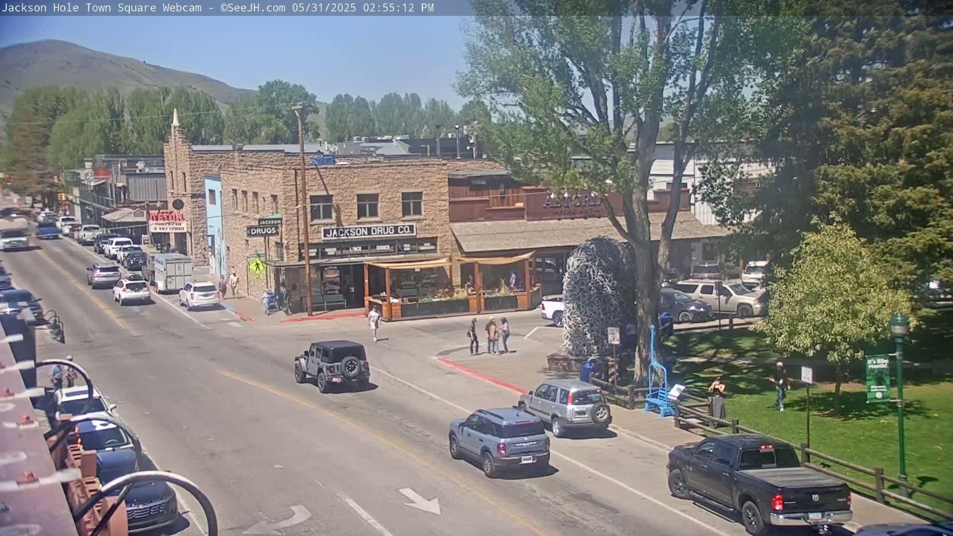 A sunny town square scene shows several cars on a street in front of various buildings, including a drug store, with people walking on the sidewalk and a grassy area with trees in the background.