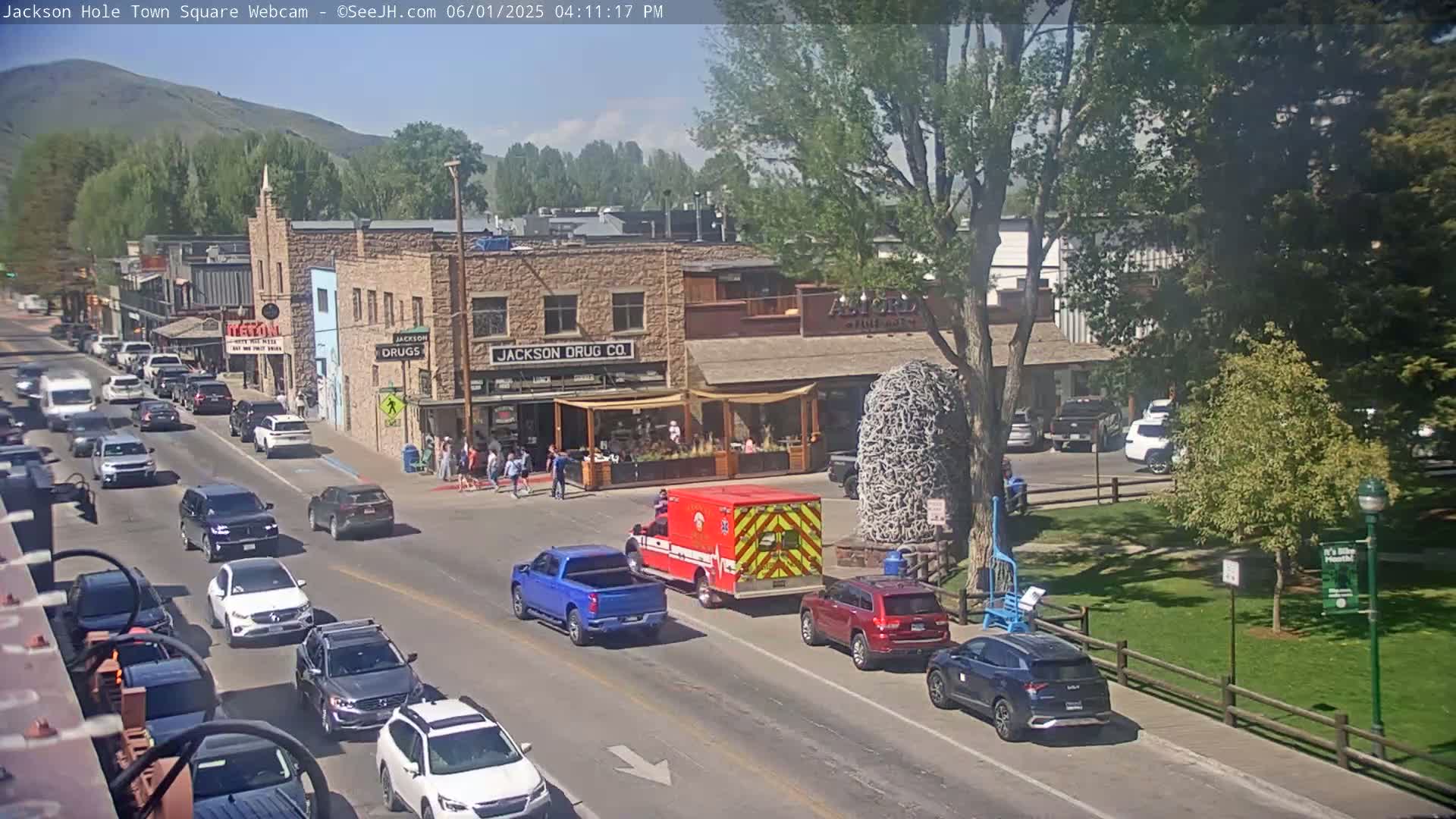 A sunny day in a town square shows several cars driving on the road near buildings, with an ambulance parked near a park.