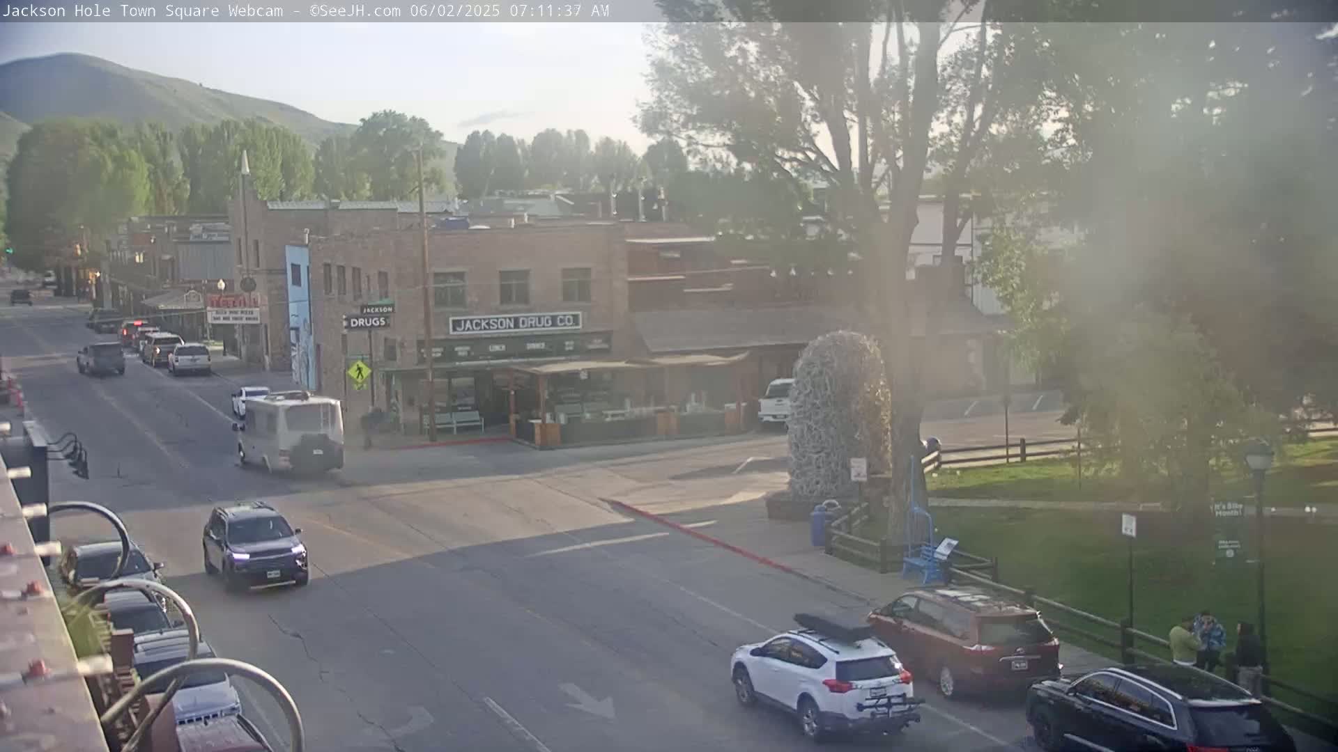 A hazy, sunny morning scene shows a town square with several vehicles driving on streets bordered by low buildings and green spaces, with trees and mountains in the distance.