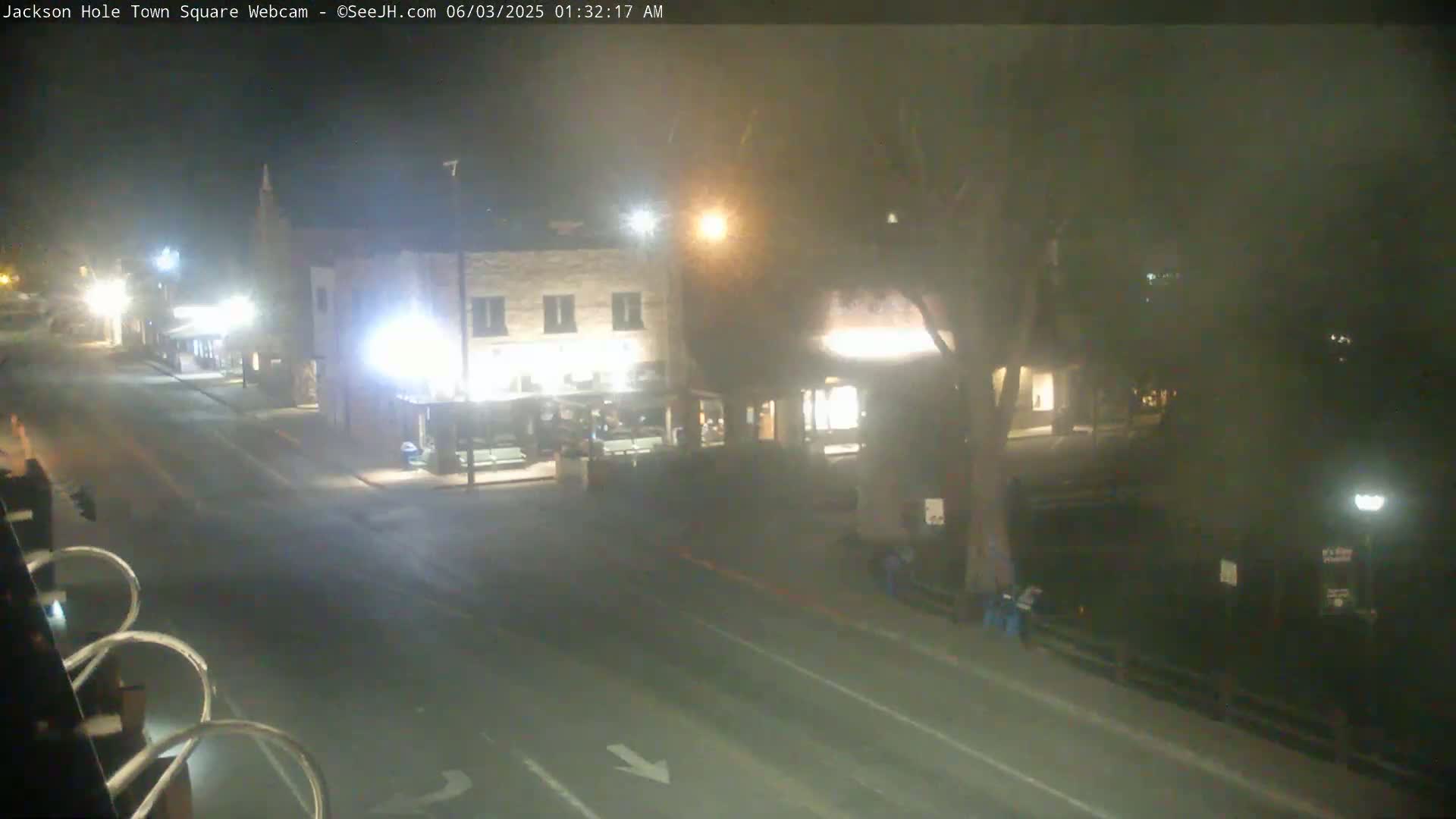 A nighttime, hazy view of a mostly empty town square with a street, buildings, and some people.