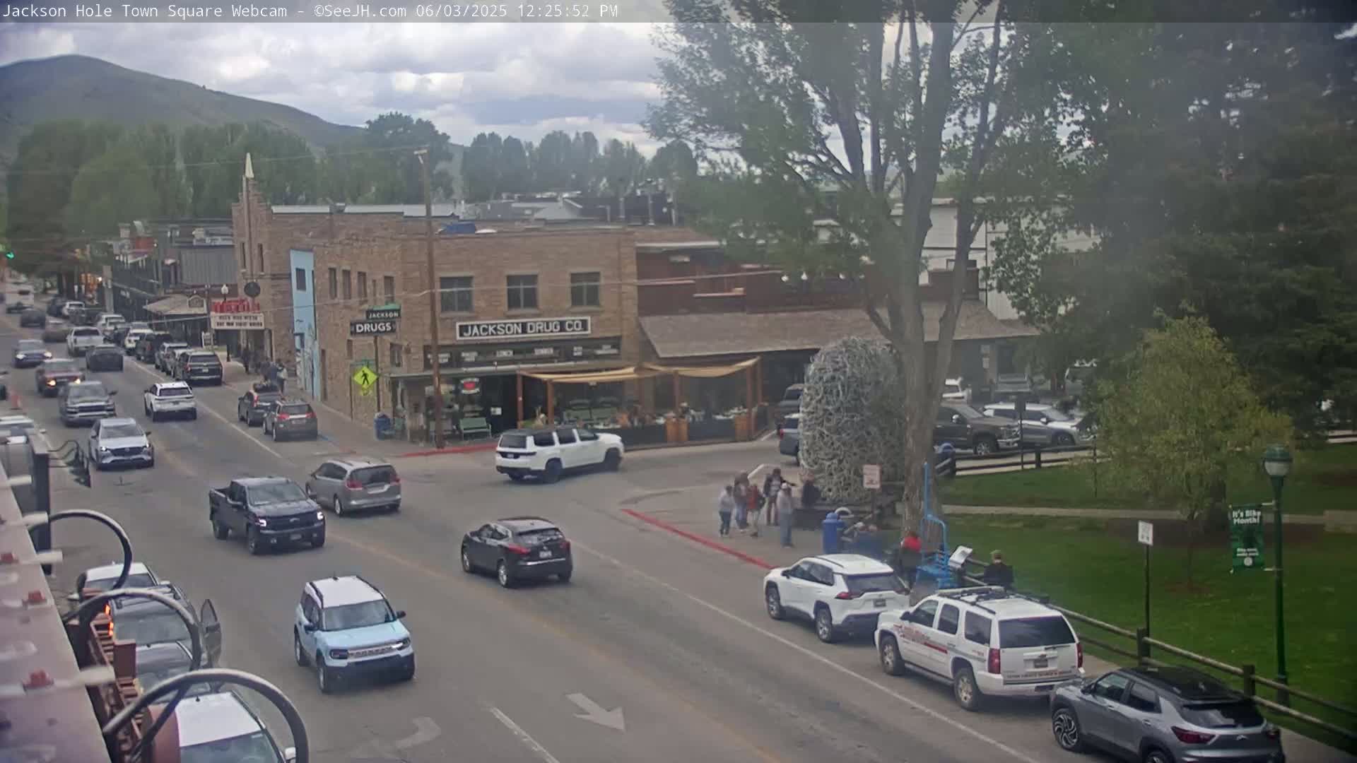 A partly cloudy day shows a town square with several cars driving on a street and people standing on a corner, with a large tree and green grass visible in the background.