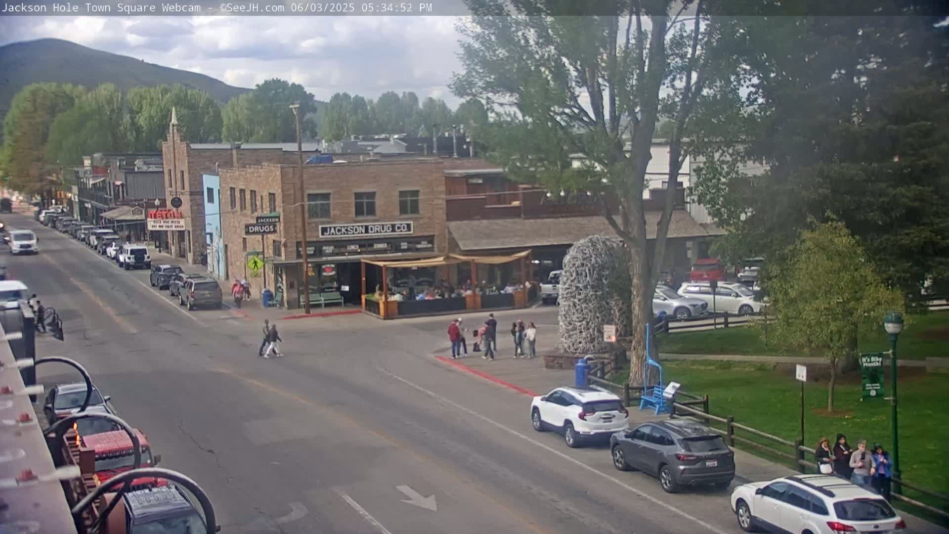 A partly cloudy day reveals a town square with parked cars, pedestrians, and buildings, including a drug store.