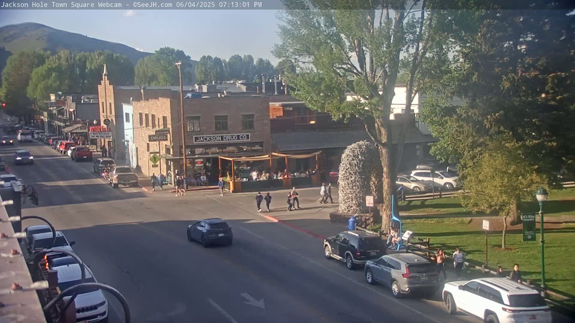 A sunny town square with cars parked along the street and pedestrians walking, under a mostly clear sky.
